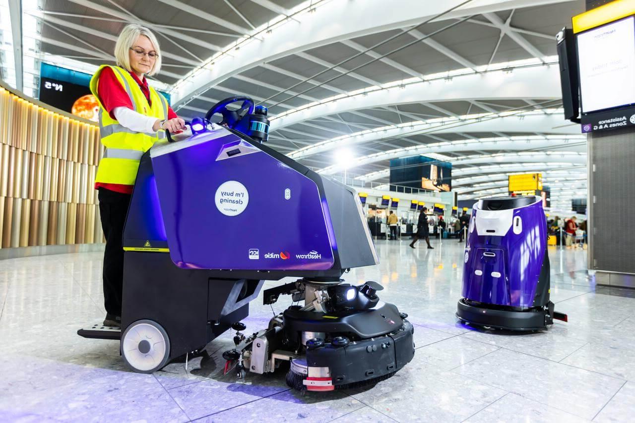 Two purple Mitie cleaning robots, with a Mitie cleaning colleague, in a London Heathrow Airport terminal
