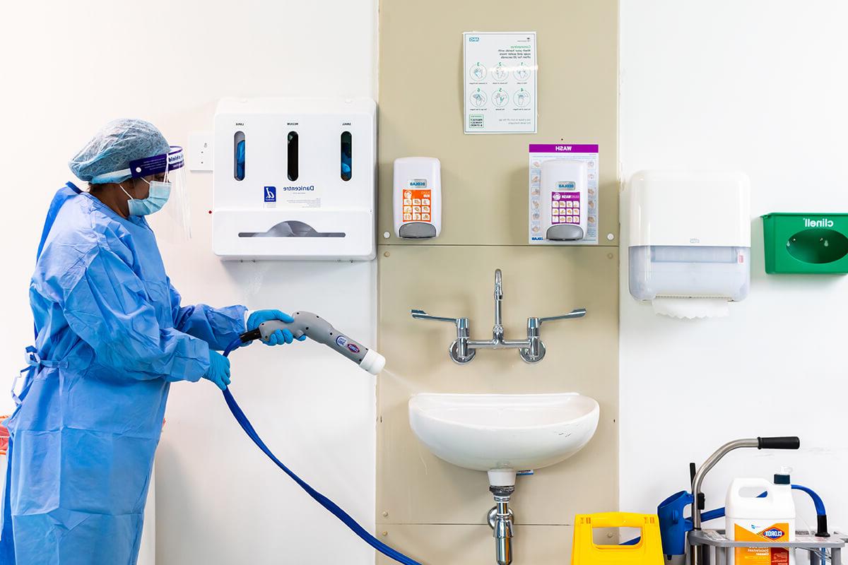A woman dressed in blue PPE is using a specialist hose to spray a white sink in a clinical setting