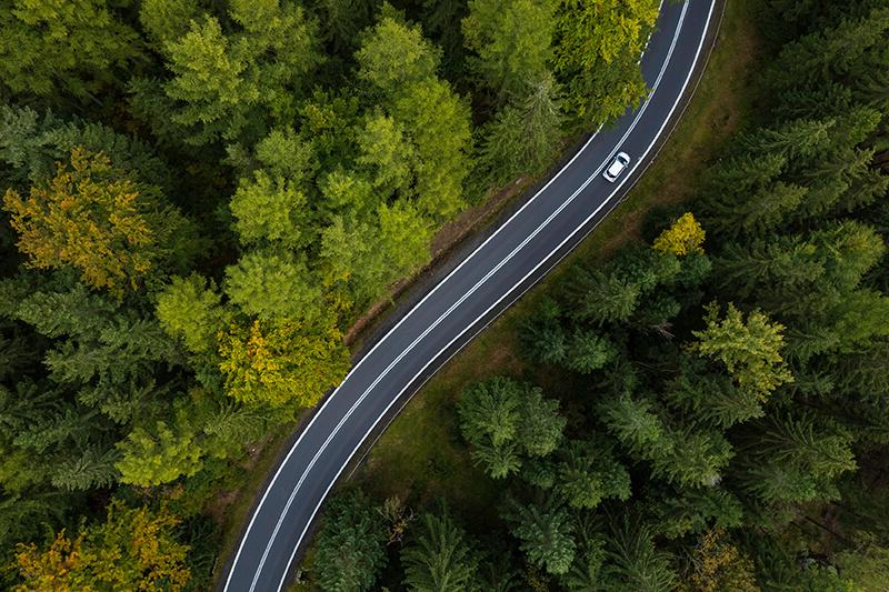Bird's eye view of a car driving on a bendy road through a surrounding green forest of trees